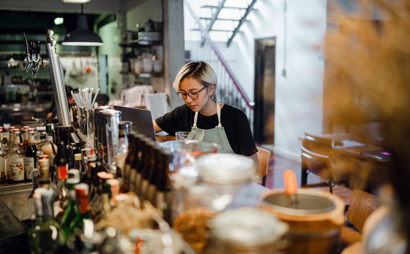 http://Focused%20female%20bartender%20using%20a%20laptop%20at%20a%20busy%20pub%20counter,%20surrounded%20by%20bottles.